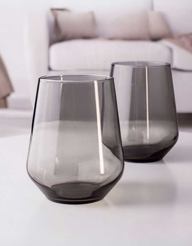 Two smoked glass tumblers on a white surface with a blurred background. Two smoky gray drinking glasses on a white table in a modern living room.